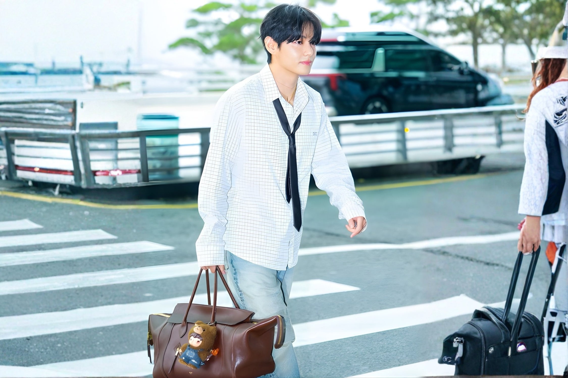 Man in casual airport style wearing checked shirt, tie, and jeans while carrying a large brown leather travel bag.