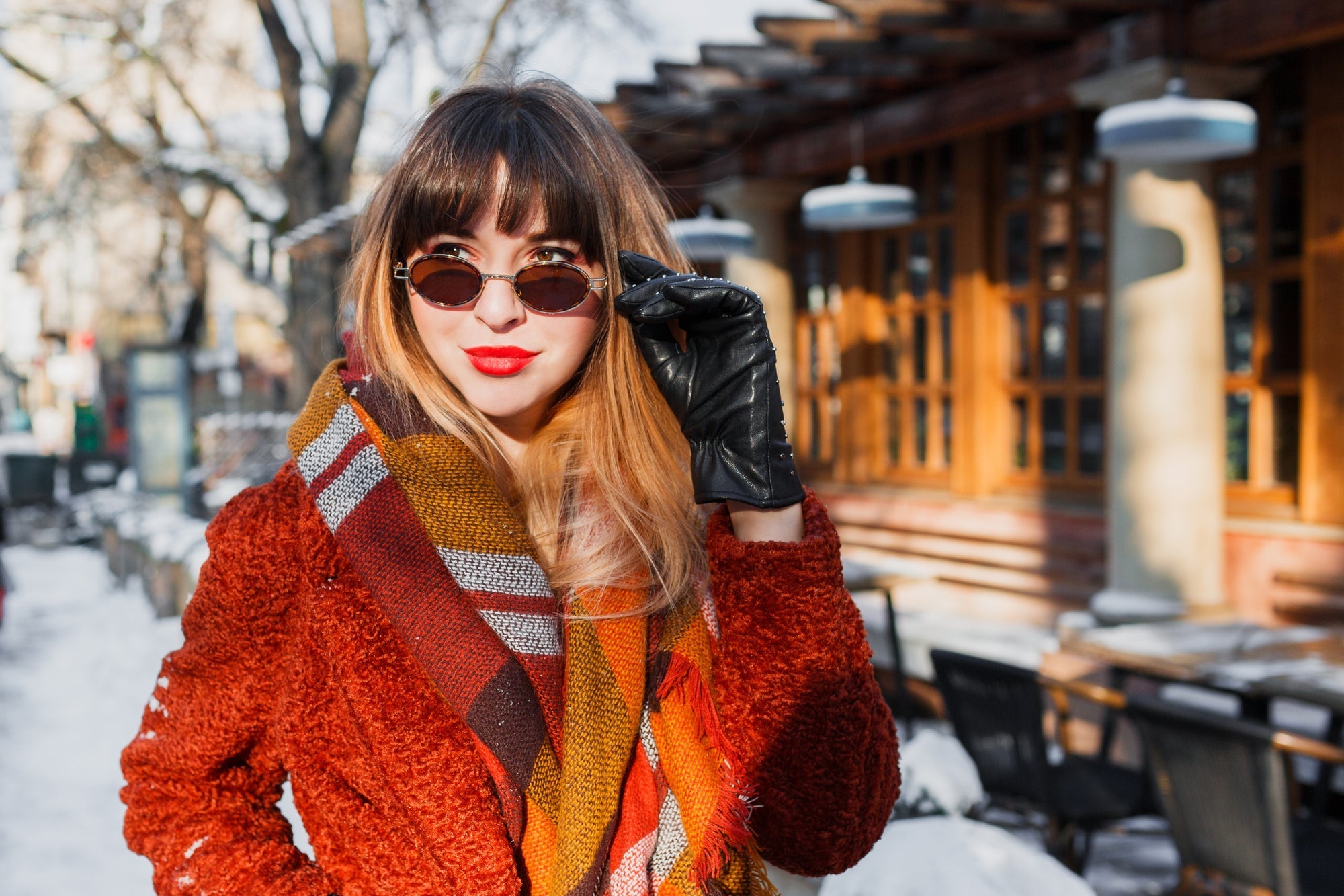 Stylish woman in a red textured coat, colorful scarf, black leather gloves, and retro sunglasses posing outdoors in a winter setting.
