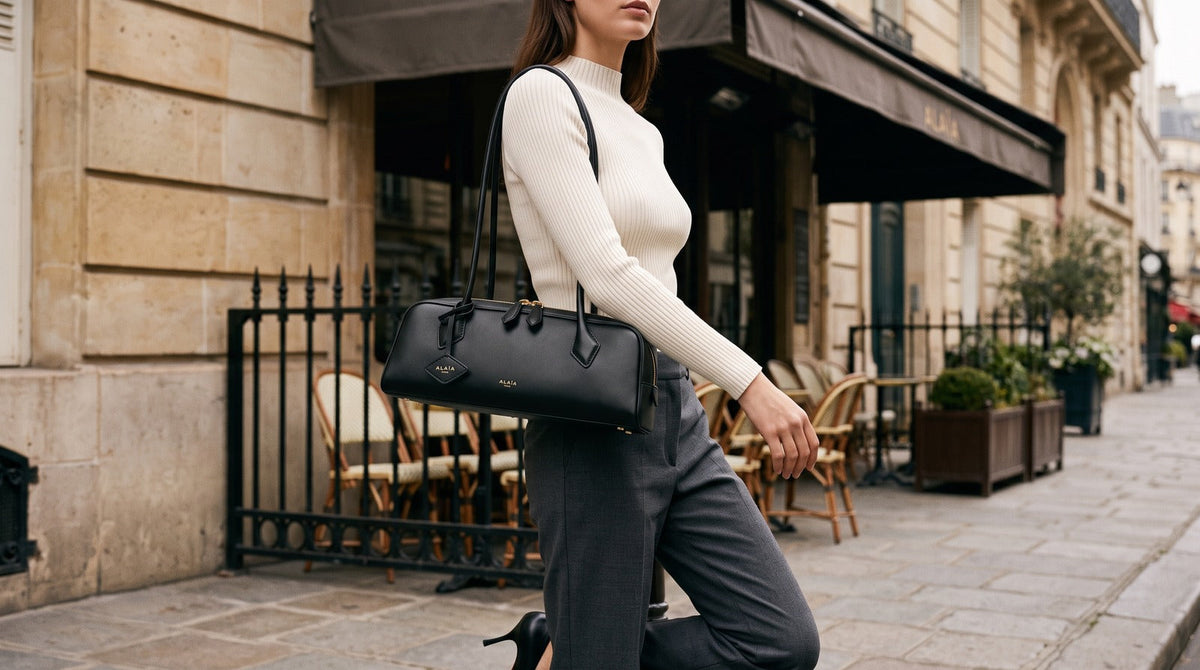 Woman carrying a black leather Alaïa Le Teckel shoulder bag, styled with ivory knitwear and charcoal trousers.