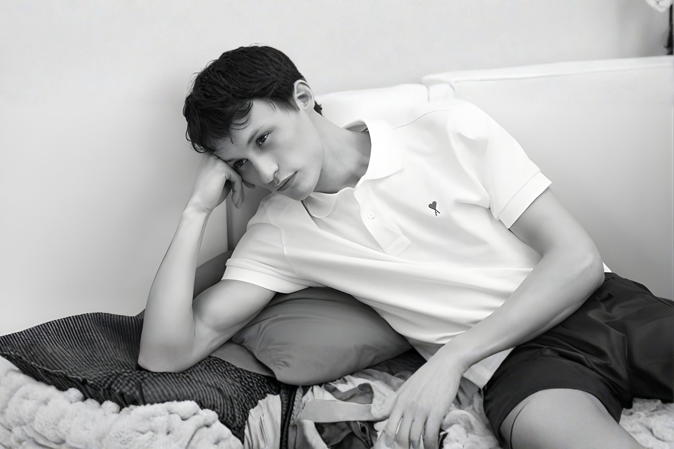 Black and white photo of a man wearing a white Ami Paris polo shirt, lounging casually on a sofa.