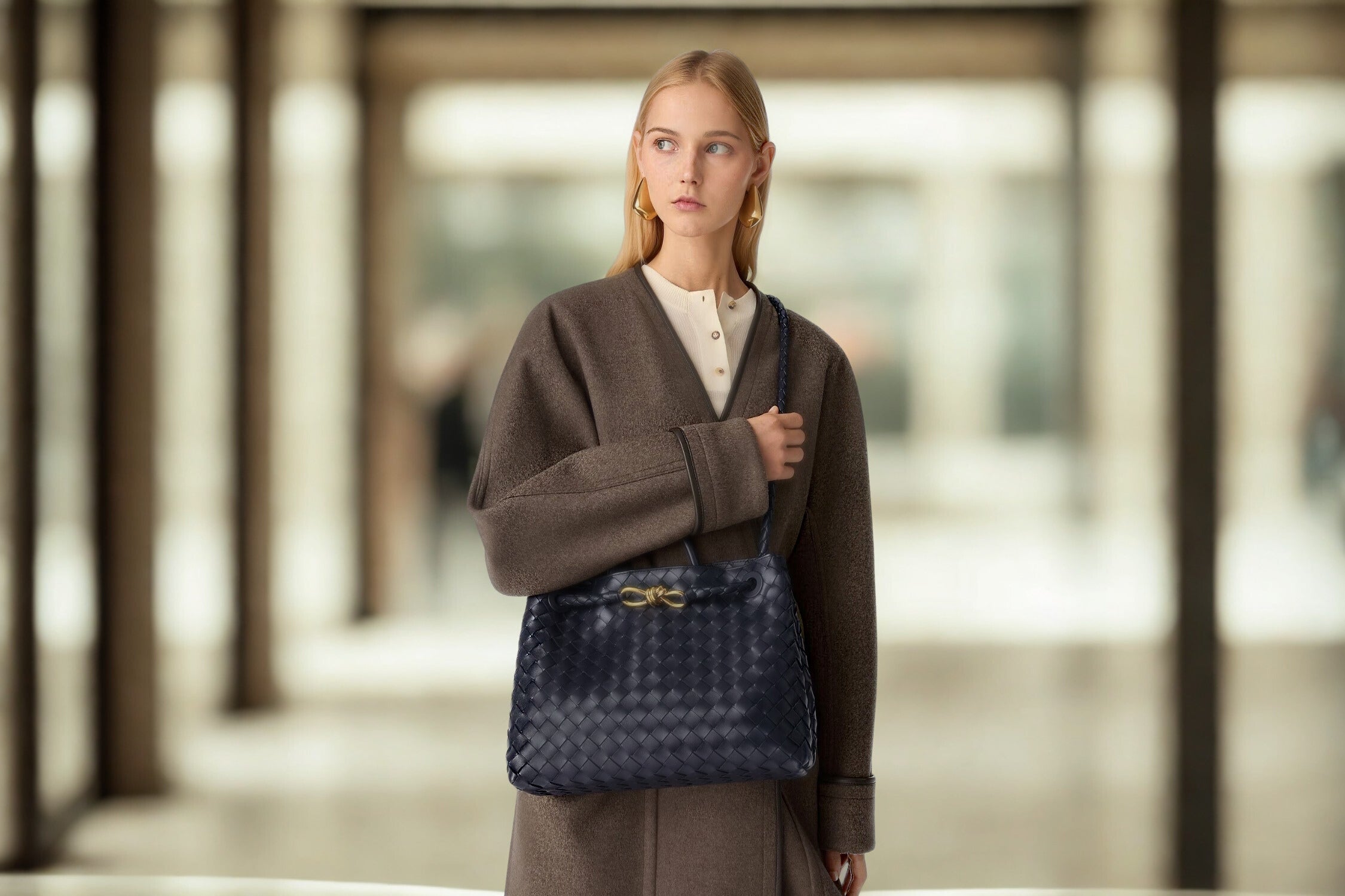Woman in a brown coat carrying a navy Bottega Veneta Andiamo bag indoors.