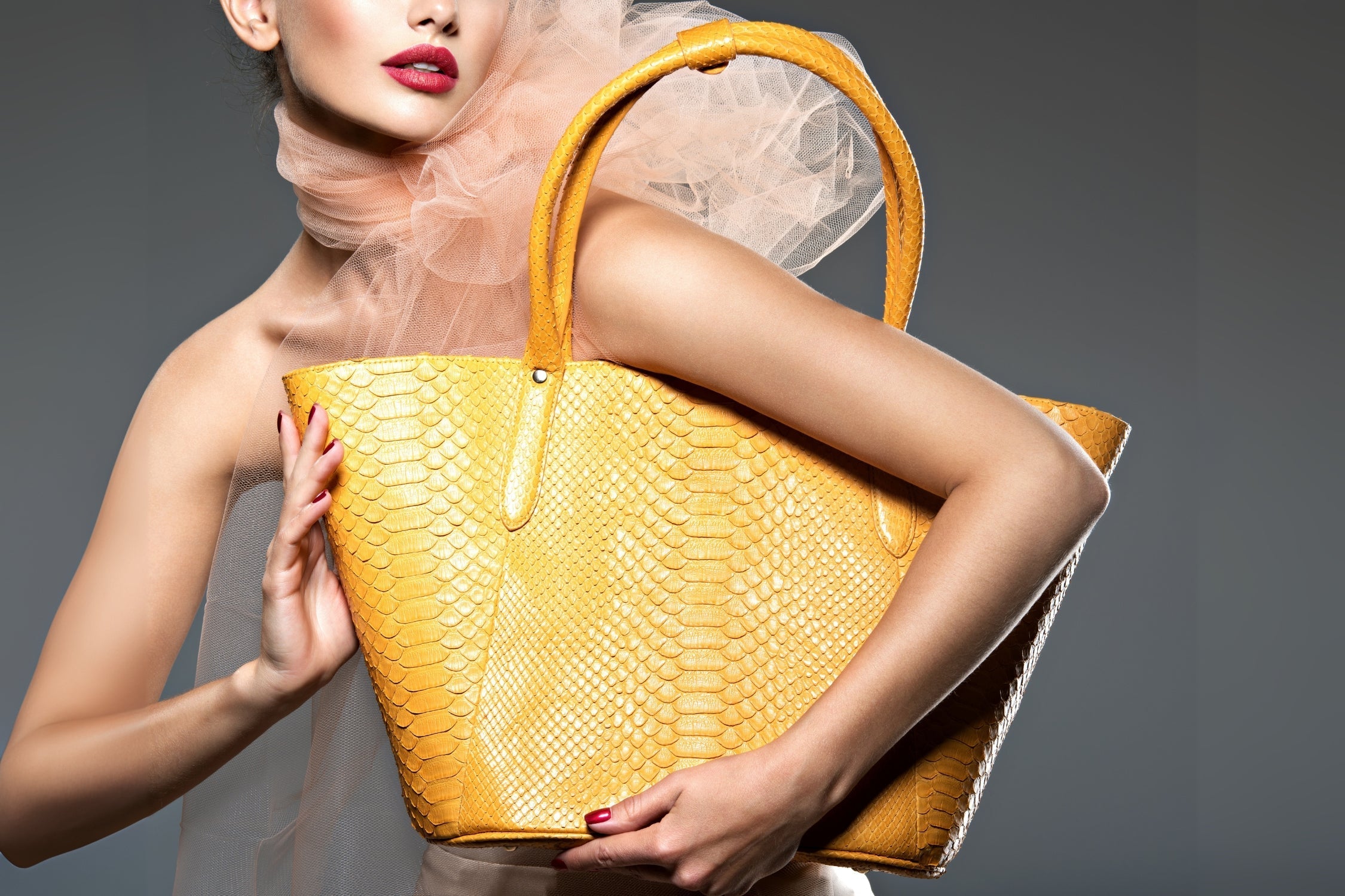 Woman holding a large yellow snakeskin handbag with elegant styling and bold red lipstick.
