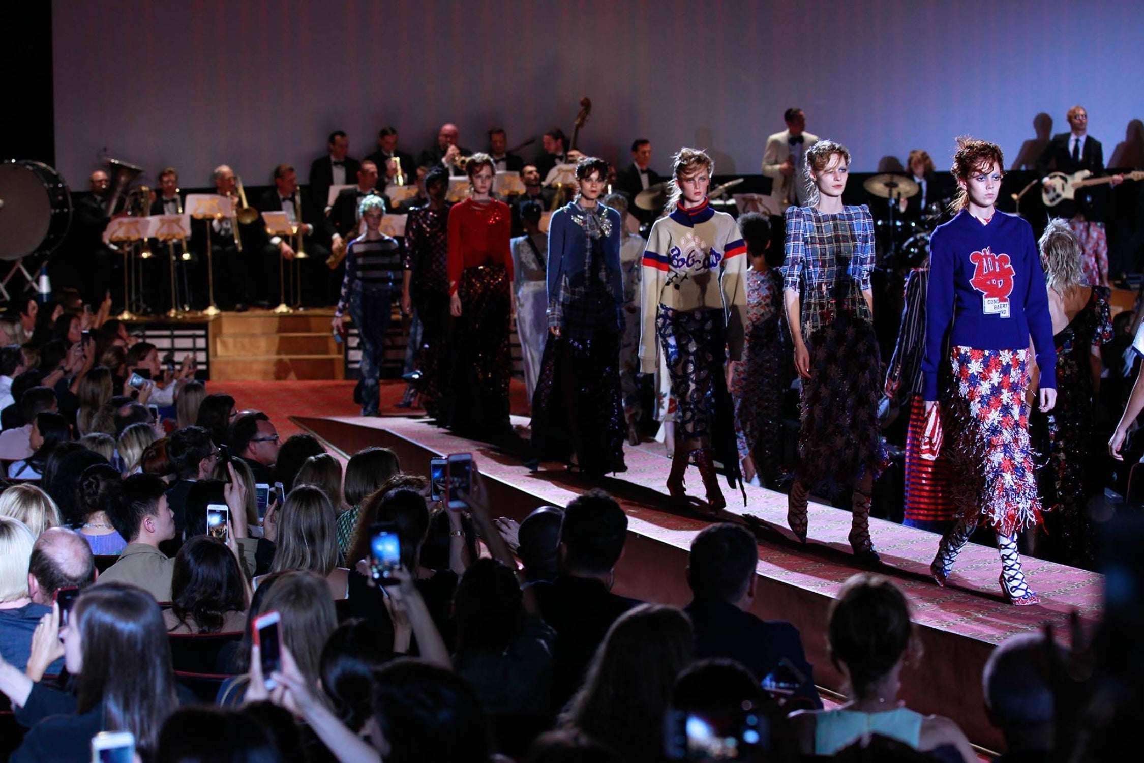 Models walking down a runway at fashion week with a live orchestra playing in the background.