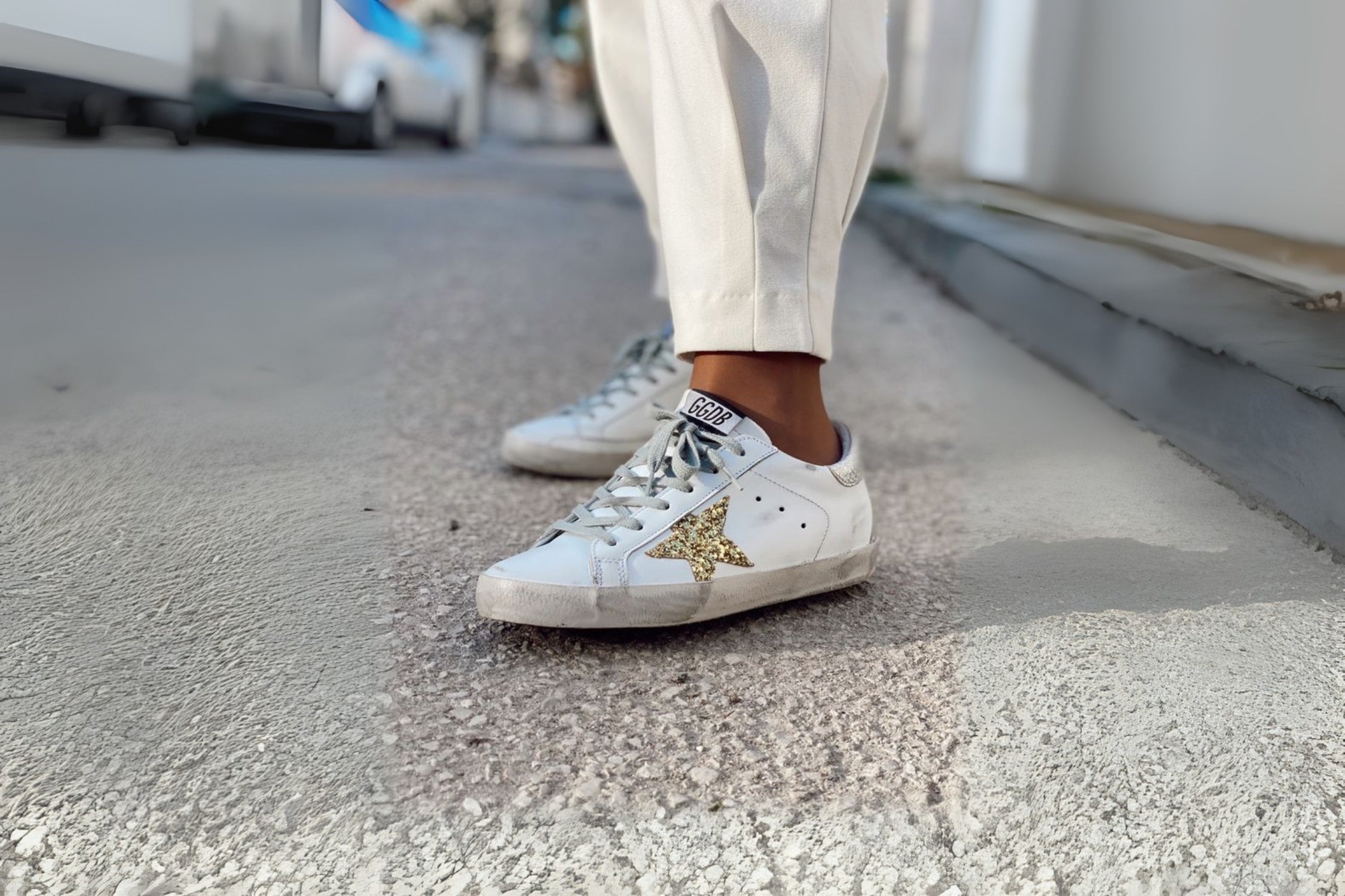 White Golden Goose sneakers with glitter star detail, worn with off-white trousers on a city sidewalk.