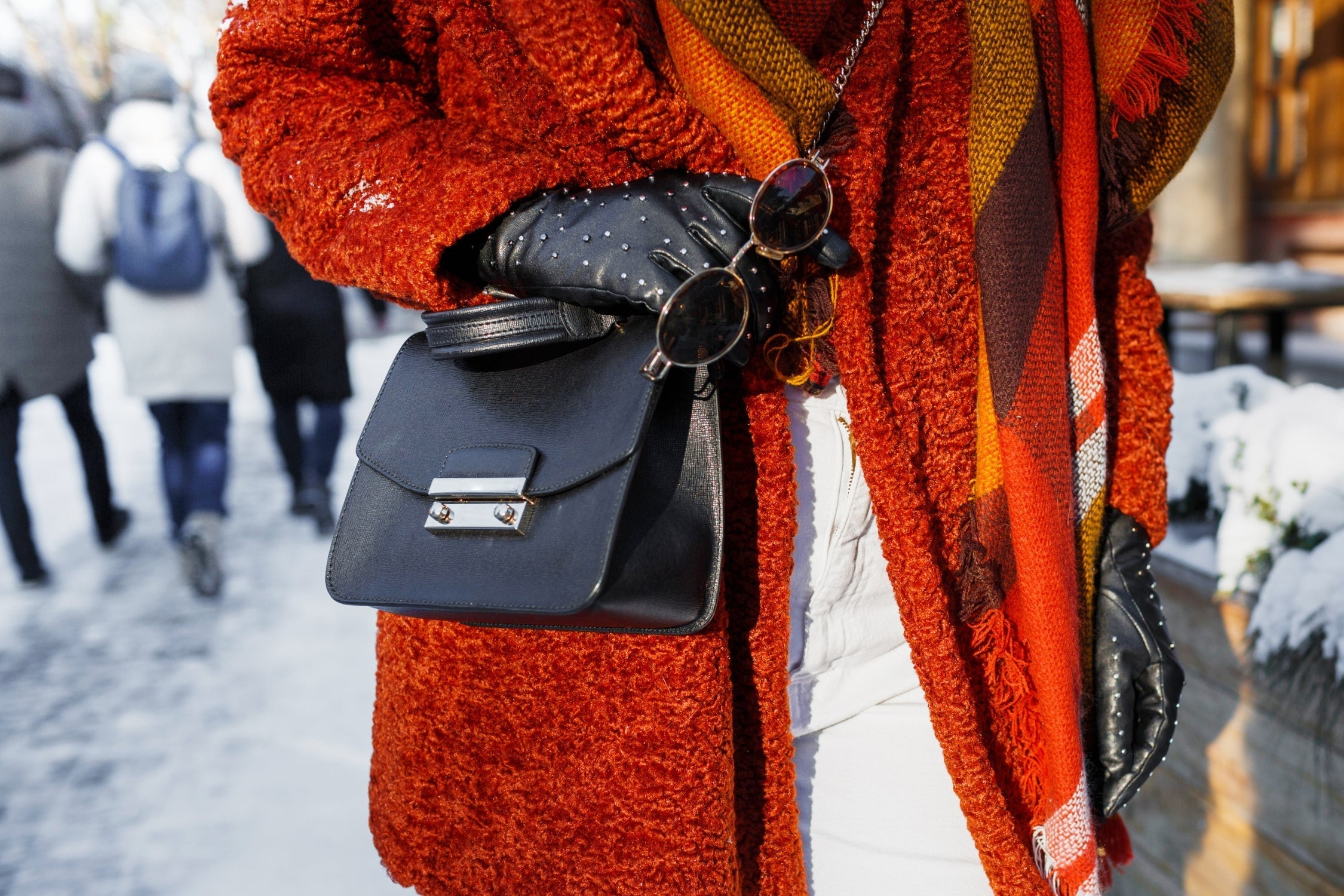 Street style close-up of a black mini handbag with silver clasp, paired with textured orange coat and leather gloves.