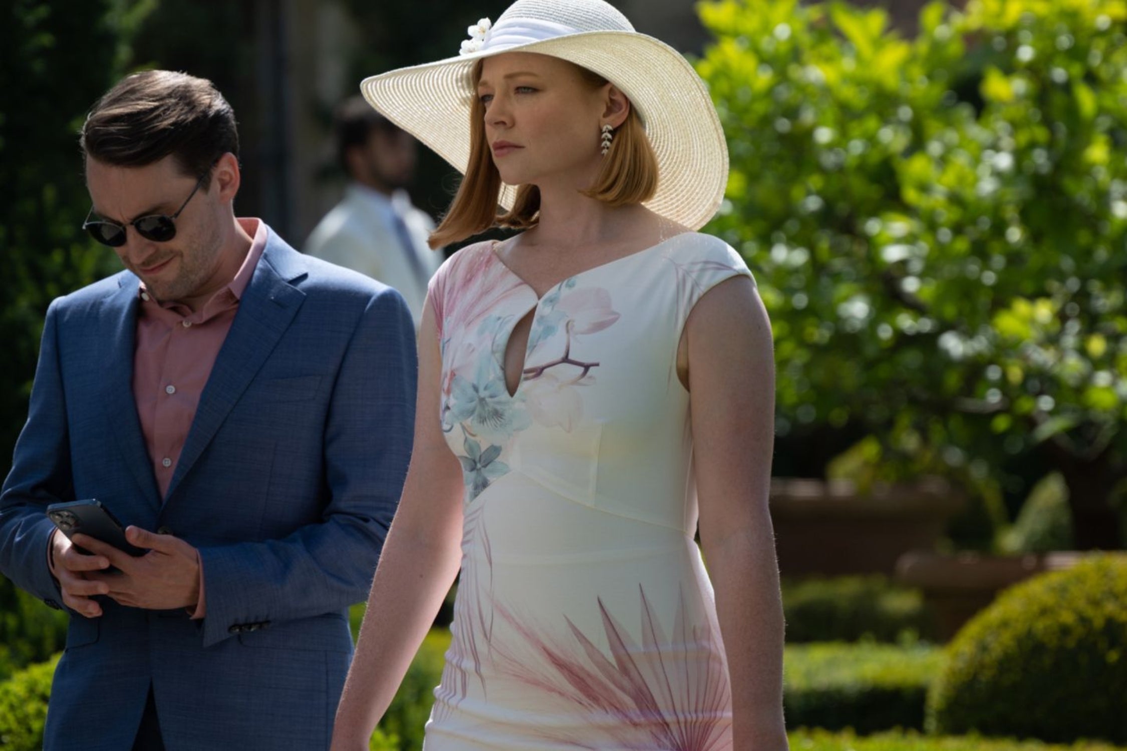 Woman in a white floral dress and wide-brim hat walking outdoors beside a man in a blue suit, set in a sunlit garden scene