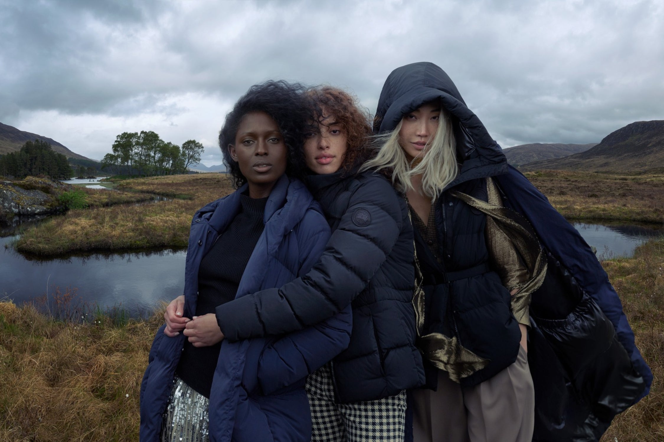 Three women wearing stylish winter outerwear standing together in a scenic, grassy landscape.