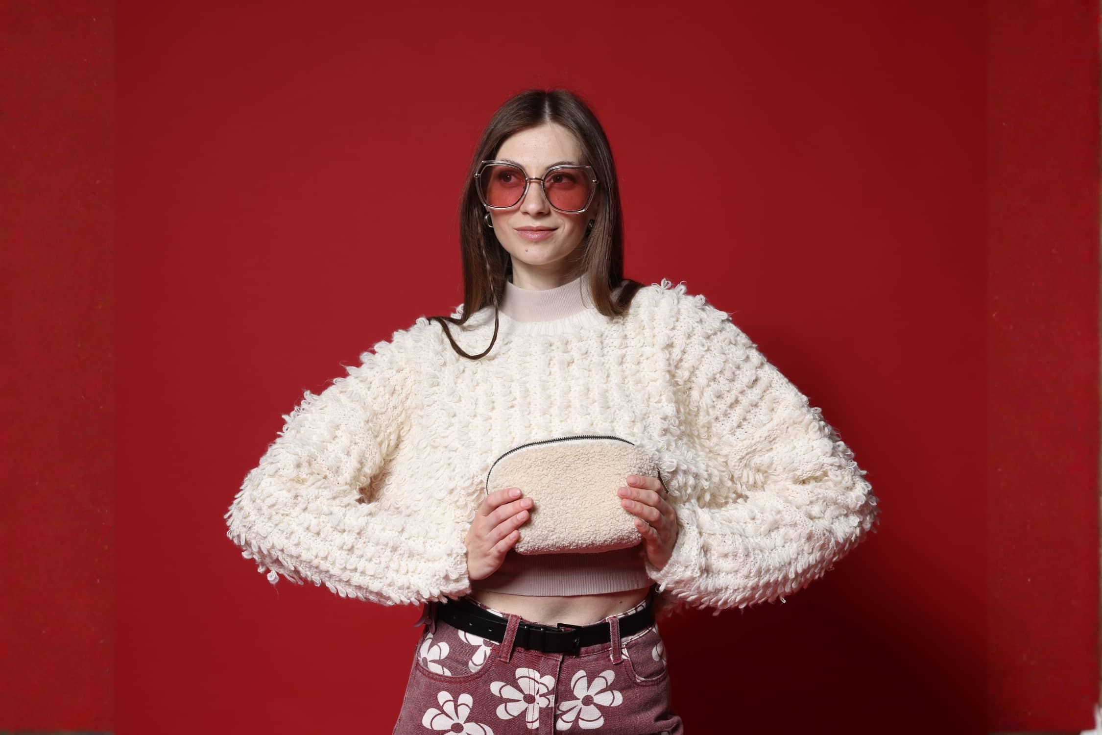 Woman wearing a cozy textured sweater for winter, holding a small beige bag against a red background.