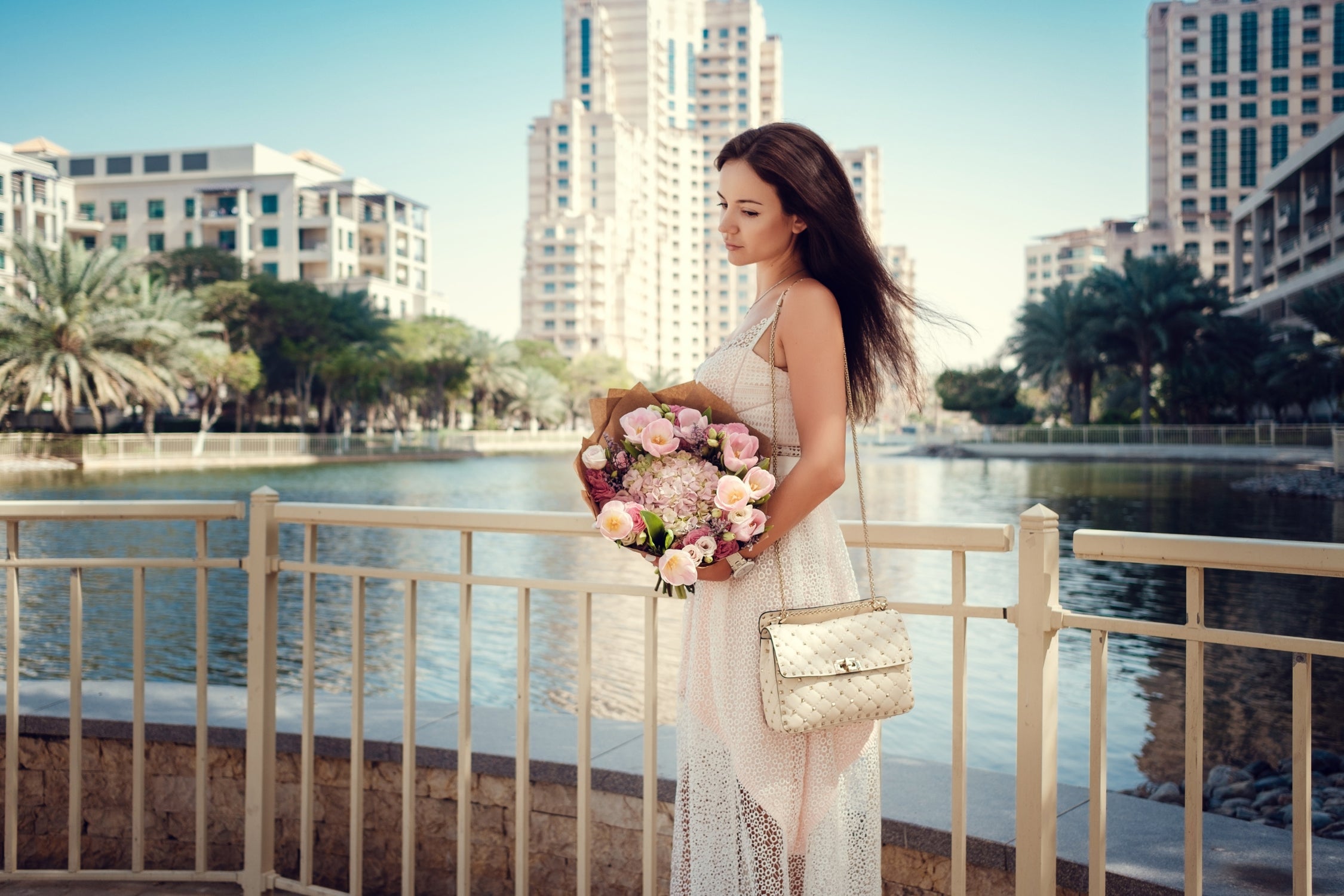 Woman in white lace dress holding bouquet by riverside, wearing quilted designer handbag.