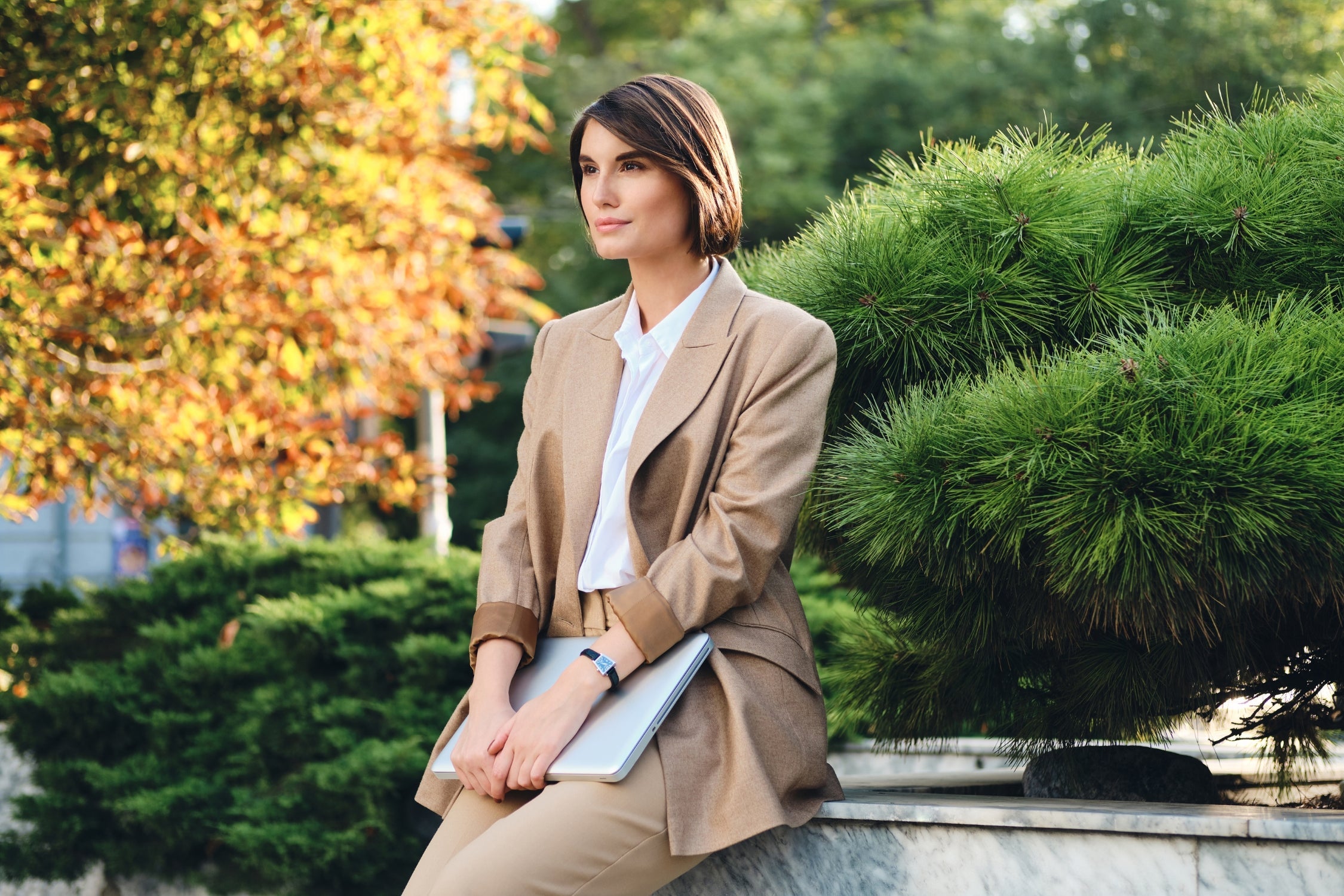 Woman in beige blazer and trousers sitting outdoors, showcasing elegant office wear style.