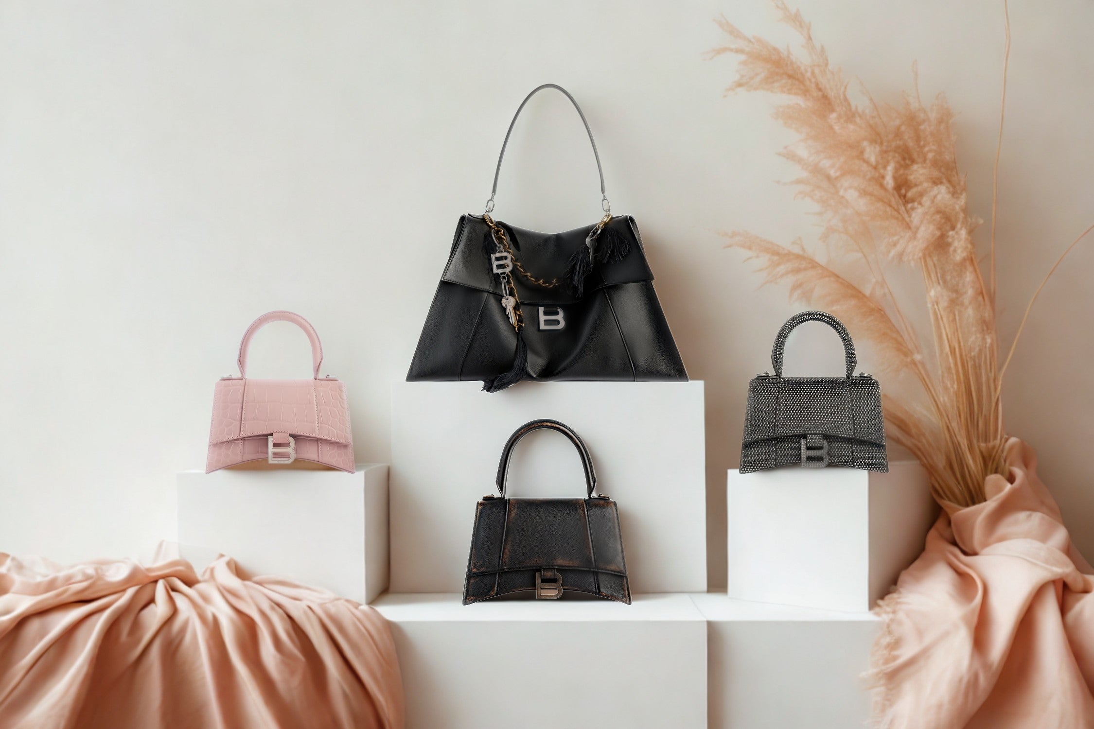 Balenciaga hourglass handbags displayed on white podiums with soft fabric and pampas grass in the background.